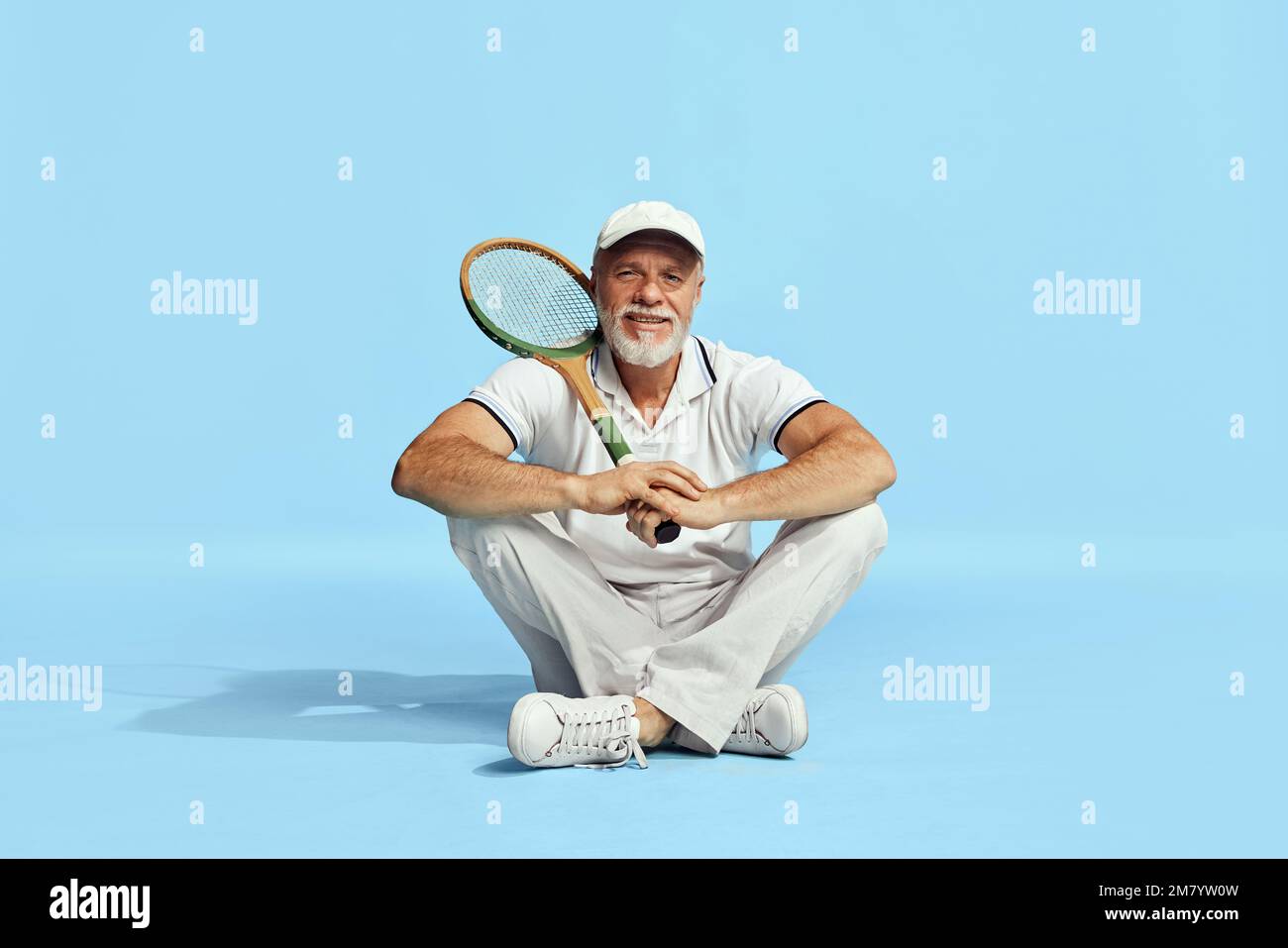 Delightful. Portrait of handsome senior man in stylish white outfit sitting, posing with tennis racket over blue background. Concept of leisure Stock Photo
