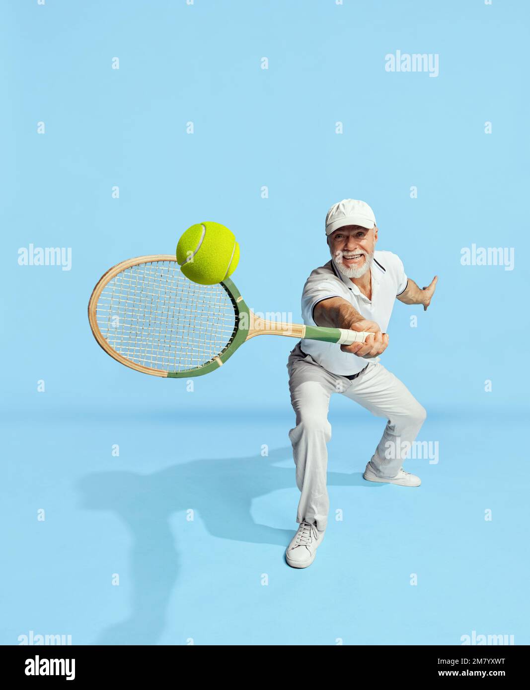 Serving ball. Portrait of handsome senior man in stylish white outfit playing tennis over blue background. Concept of leisure activity, hobby Stock Photo