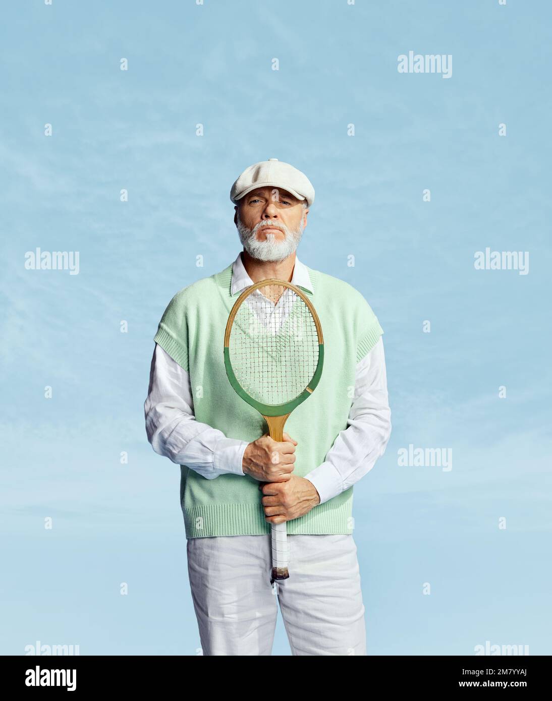 Serious look. Portrait of handsome senior man in stylish white outfit posing with tennis racket over sky blue background. Concept of leisure activity Stock Photo