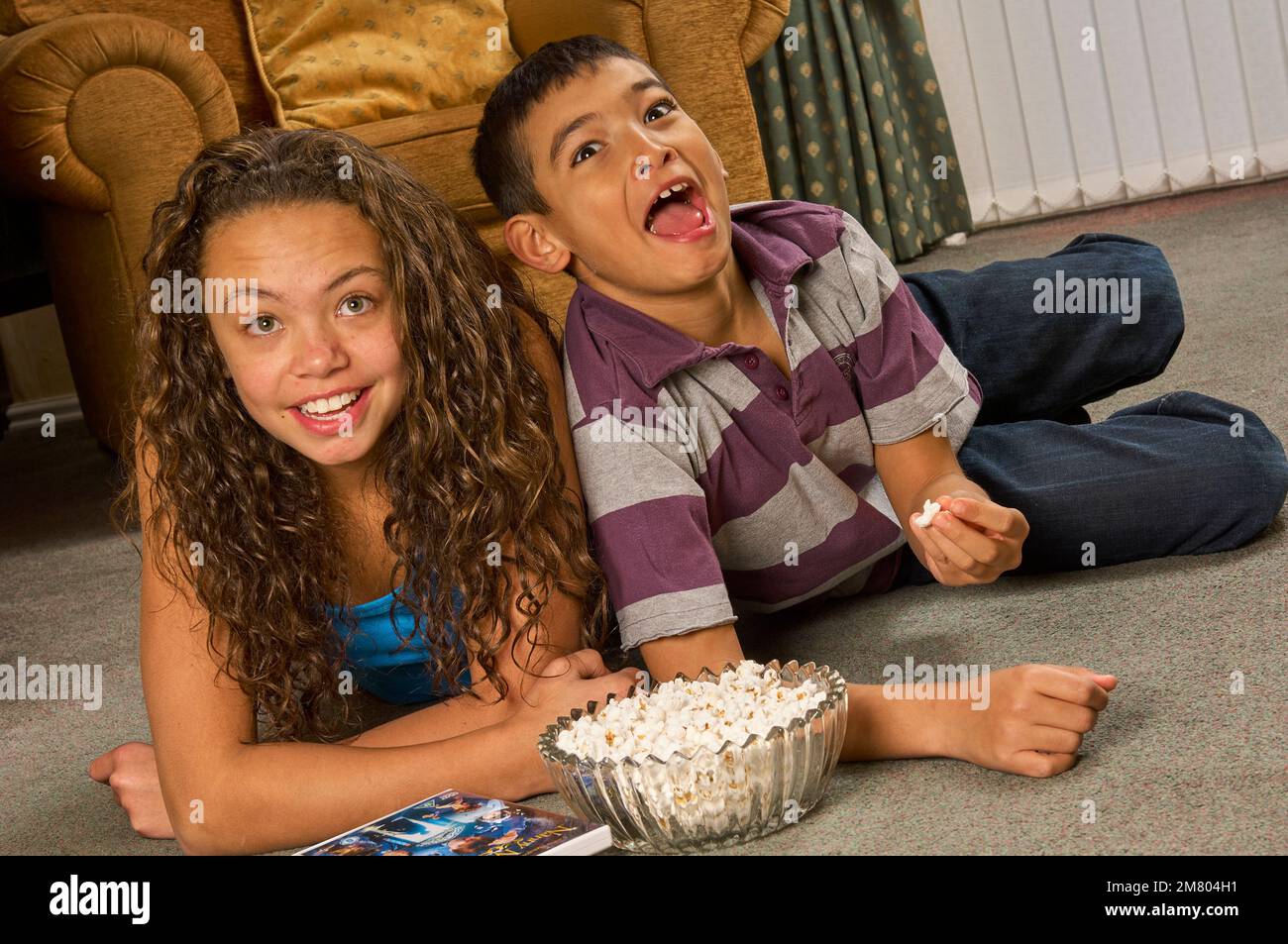 Young teenage girl and young boy watching a film together lying on the floor in a lounge at home, eating popcorn Stock Photo