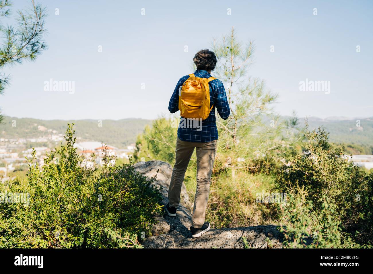 Man hiker with yellow backpack standing on the mountain rock. A tourist stands against the background of a valley. Rearview of a male hiker standing Stock Photo