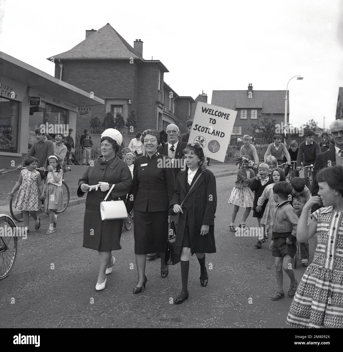 1960s, historical, excited children in the street, as a lady from the Salvation Army in Canada pays a vistor to the twon/village, Fife, Scotland, UK. Man carries a placard, saying 'Welcome to Scotland', Maple leaf and thistles on it Stock Photo