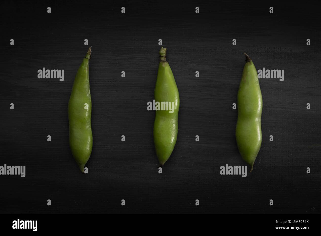three broad beans in shell lined up on a black wooden background. Stock Photo