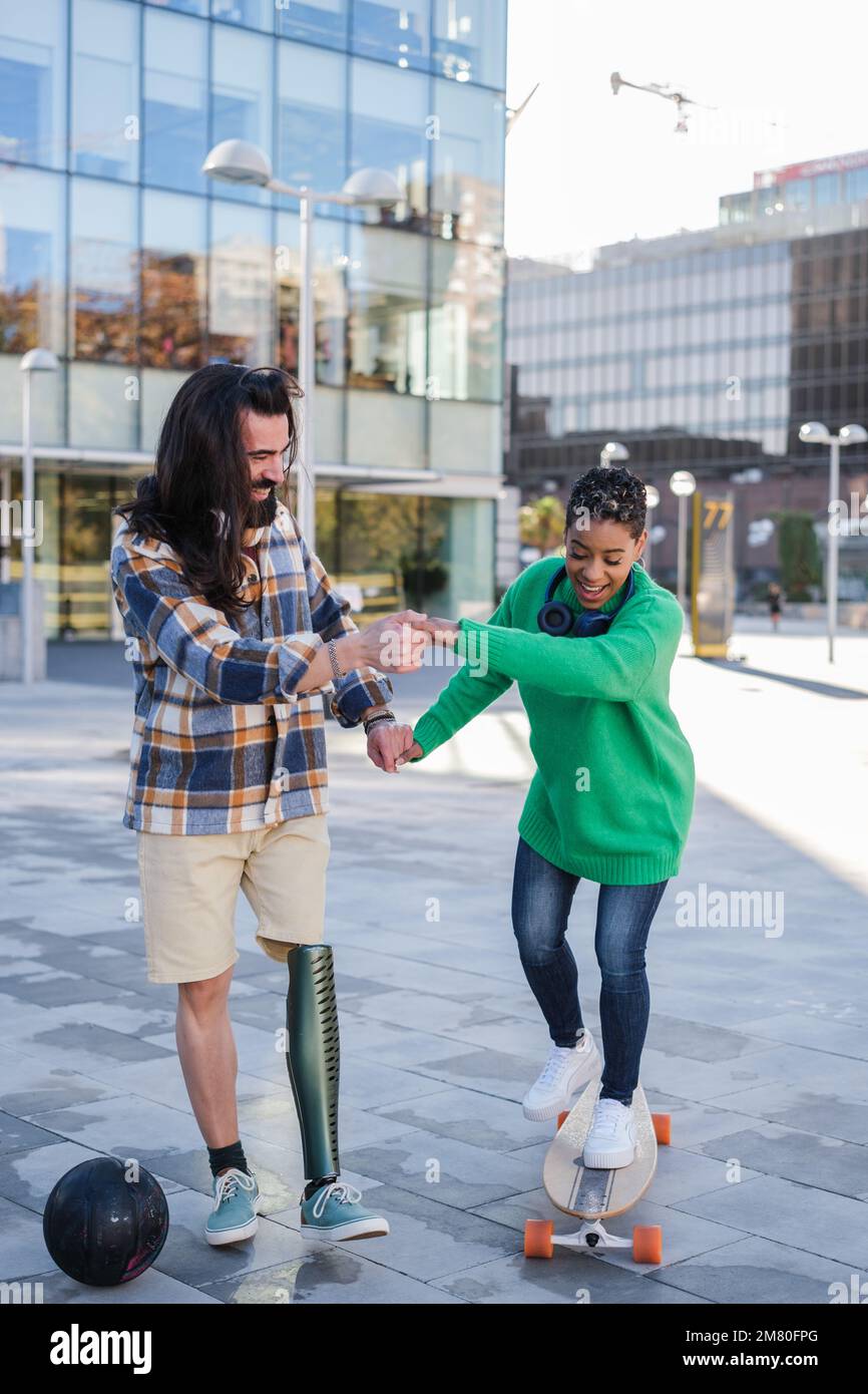 Young man with prosthetic leg teaching his girlfriend to ride a skateboard outdoors - learning and improvement concept Stock Photo