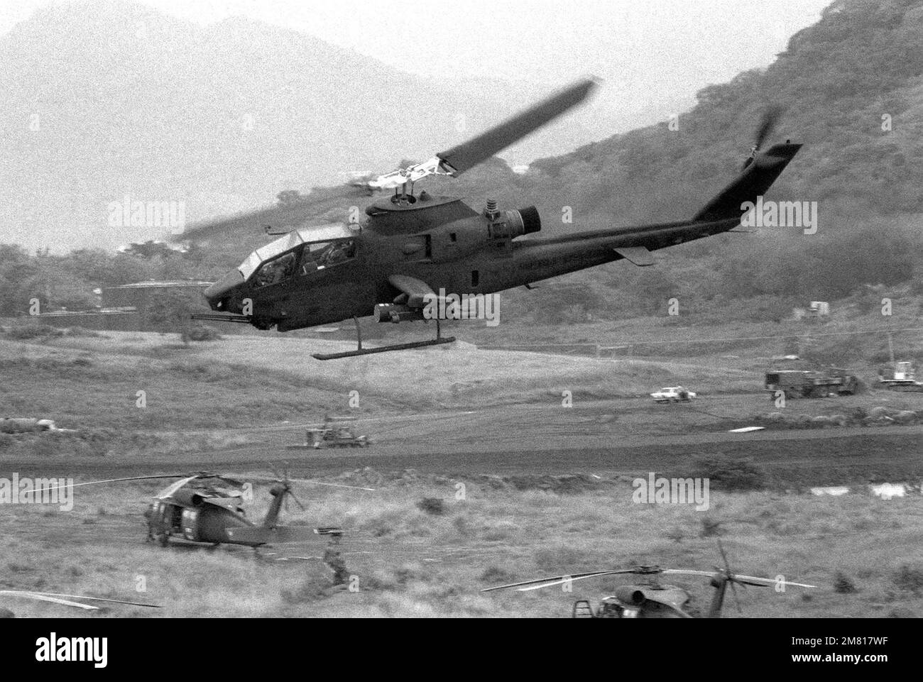 An AH-1 Cobra helicopter flies over several UH-60 Black Hawk (Blackhawk) helicopters parked in a field at Point Salines Airfield during the multiservice, multinational Operation Urgent Fury. Subject Operation/Series: URGENT FURY Country: Grenada (GRD) Stock Photo