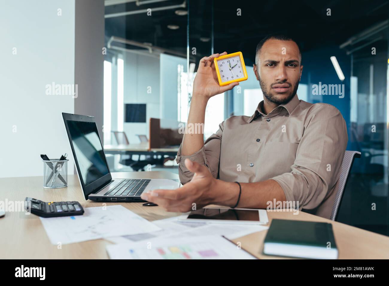 Dissatisfied hispanic man looking at camera and showing clock, businessman frustrated waiting for colleague to be late, working inside office with documents using laptop inside office. Stock Photo