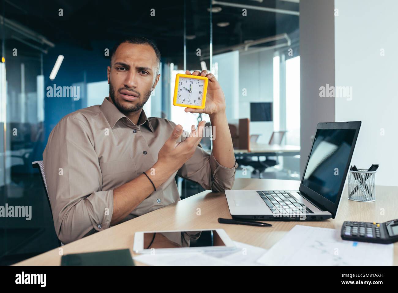 Dissatisfied hispanic man looking at camera and showing clock, businessman frustrated waiting for colleague to be late, working inside office with documents using laptop inside office. Stock Photo