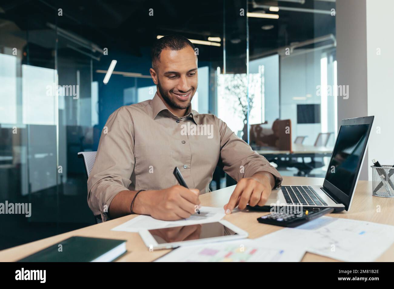 Successful african american businessman inside office doing paperwork, man writing information, worker using laptop for work sitting with bills and contracts smiling and satisfied with result. Stock Photo