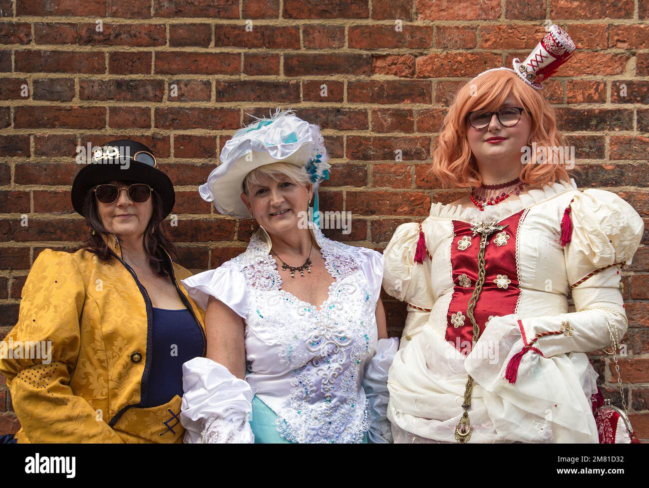 Portrait of three women, female steampunks, smiling while standing next to a wall. Dressed in period costume, steampunk clothing. Stock Photo