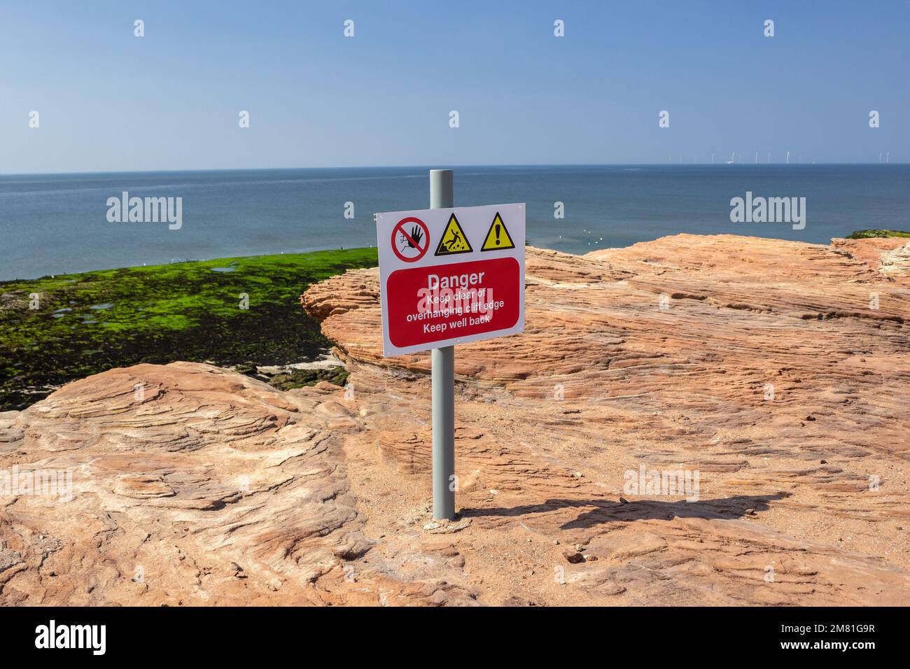 Hilbre Island, UK: Warning sign on rocky ledge with the Dee estuary below. Danger - Keep clear of overhanging cliff edge, Keep well back Stock Photo