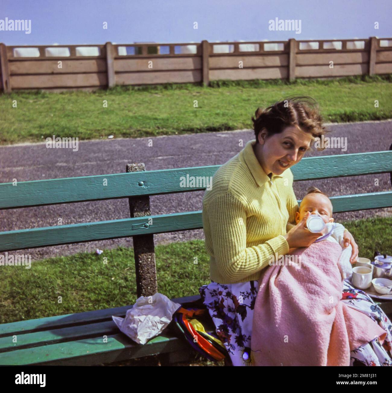 A young mother bottle feeds her baby in 1957. She is sitting on a wooden seat near the coast at Great Yarmouth, England. This photo was taken from the original slide. Stock Photo