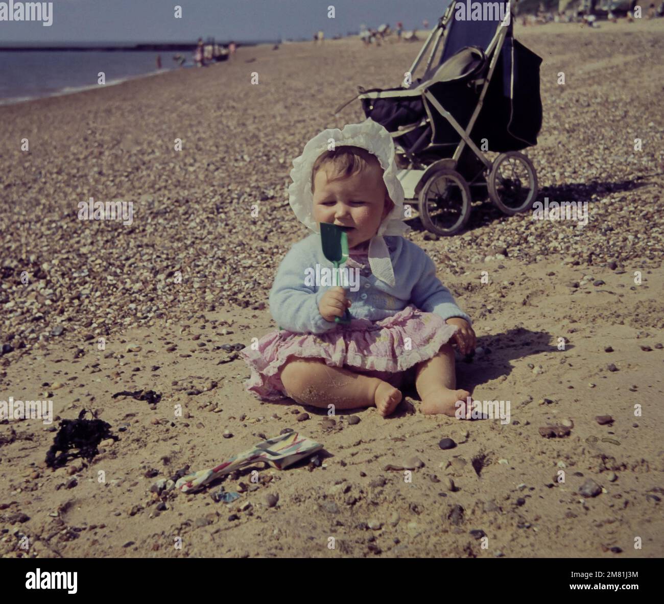 A one year old girl plays with a spade on Clacton Beach in Essex in 1960. The girl wears a white cotton bonnet to protect her from the sun. The photo is taken from the original slide. Stock Photo