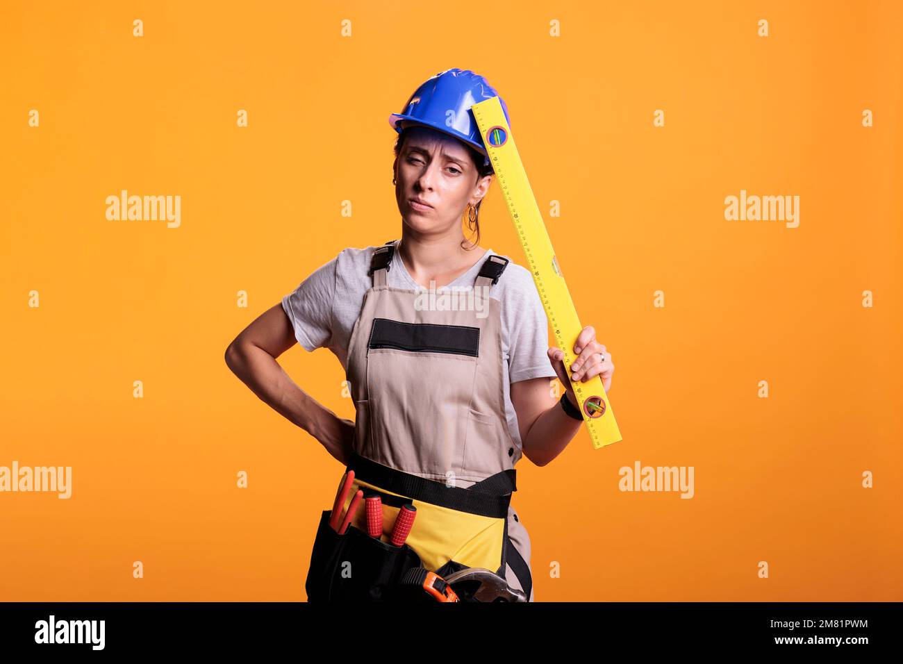 Construction worker suffering from painful headache and holding leveler on camera, rubbing temples. Woman renovation expert with migraine dressed with overalls and hardhat in studio. Stock Photo