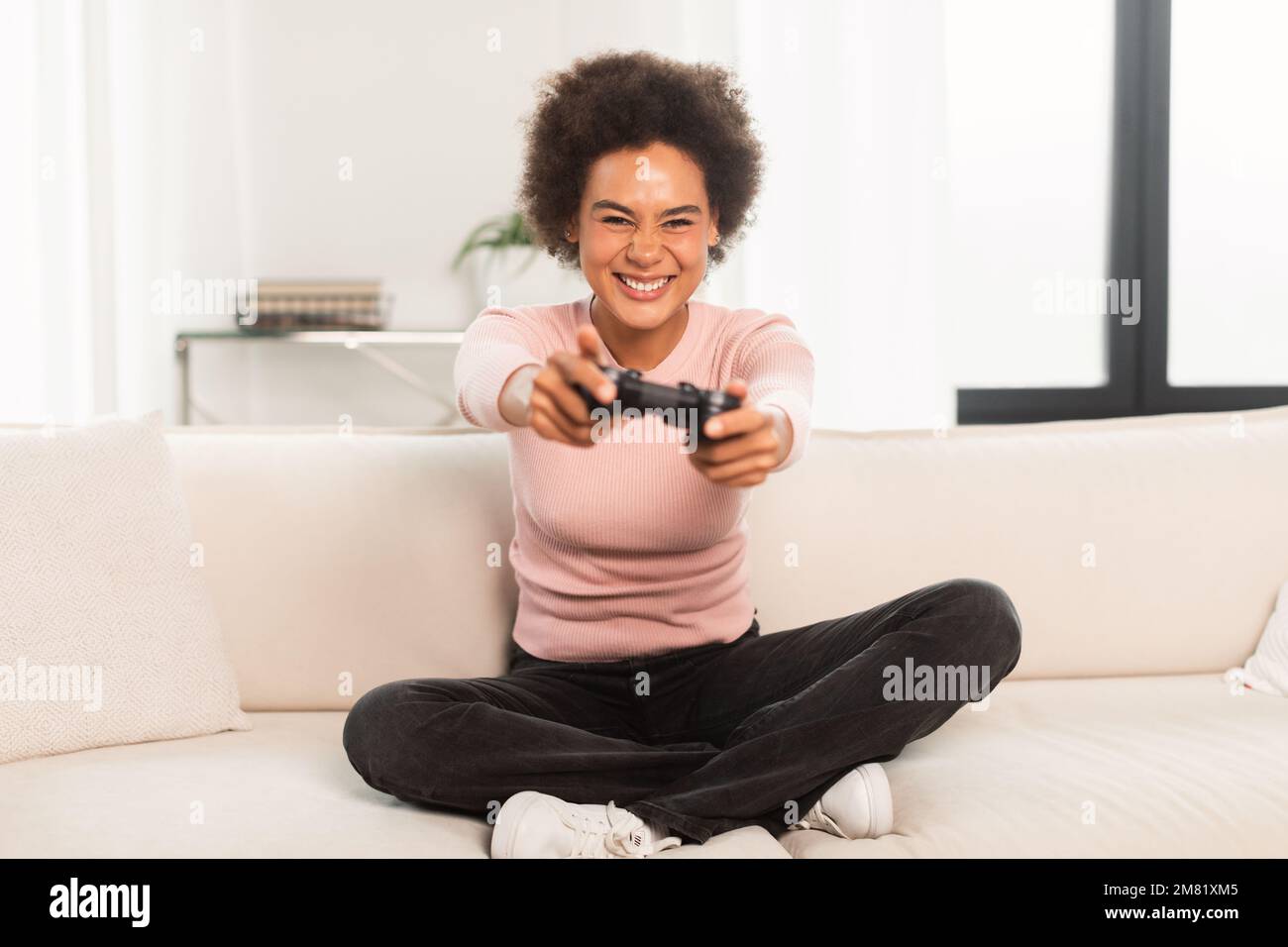 Cheerful excited young mixed race woman sitting on sofa with joystick, playing computer game in living room Stock Photo