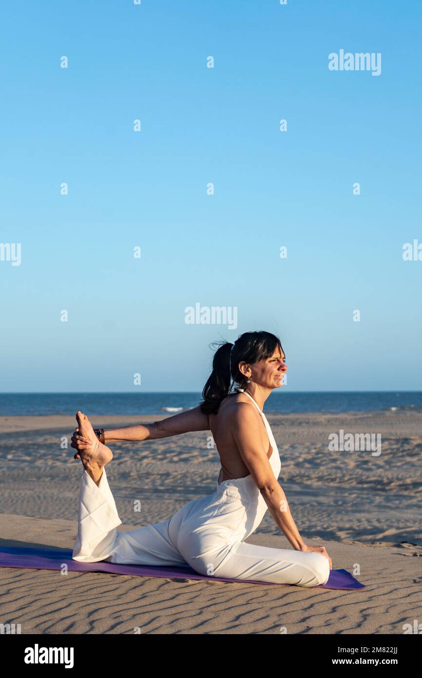 A woman practicing yoga on a mat on the beach next to the ocean Stock Photo