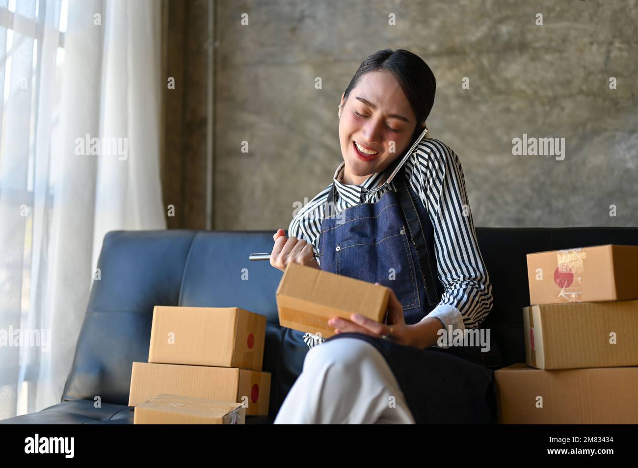 Charming and busy millennial Asian female e-commerce business entrepreneur or online seller is on the phone with her suppliers while preparing shippin Stock Photo