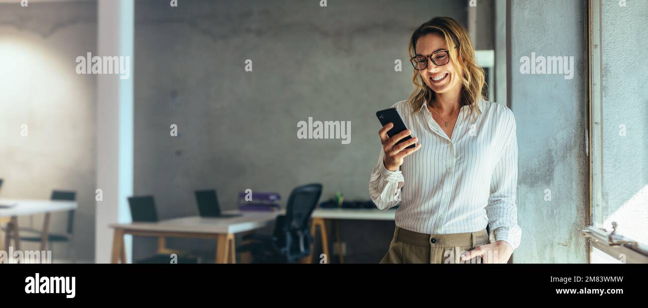 Smiling businesswoman using her phone in the office. Small business entrepreneur looking at her mobile phone and smiling while communicating with her Stock Photo