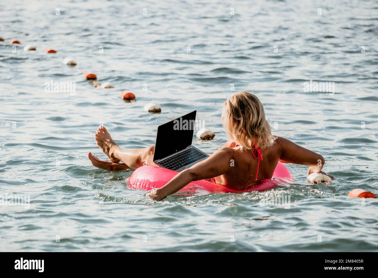 Woman works on laptop in sea. Freelancer, young blond woman in sunglases floating on an inflatable big pink donut with a laptop in the sea at sunset Stock Photo