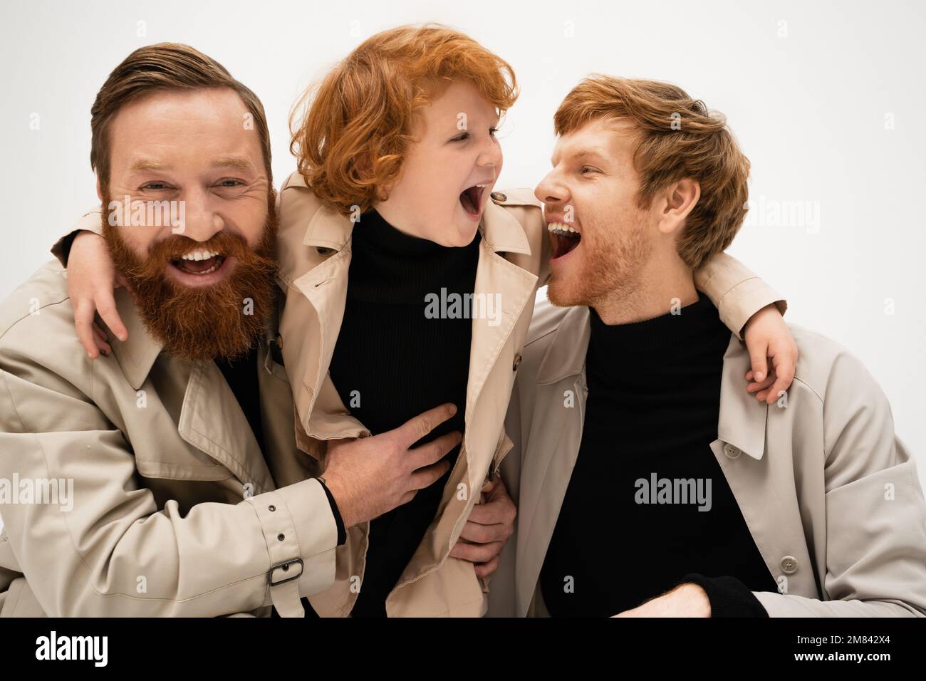 joyful bearded men and red haired boy in trench coats laughing isolated ...