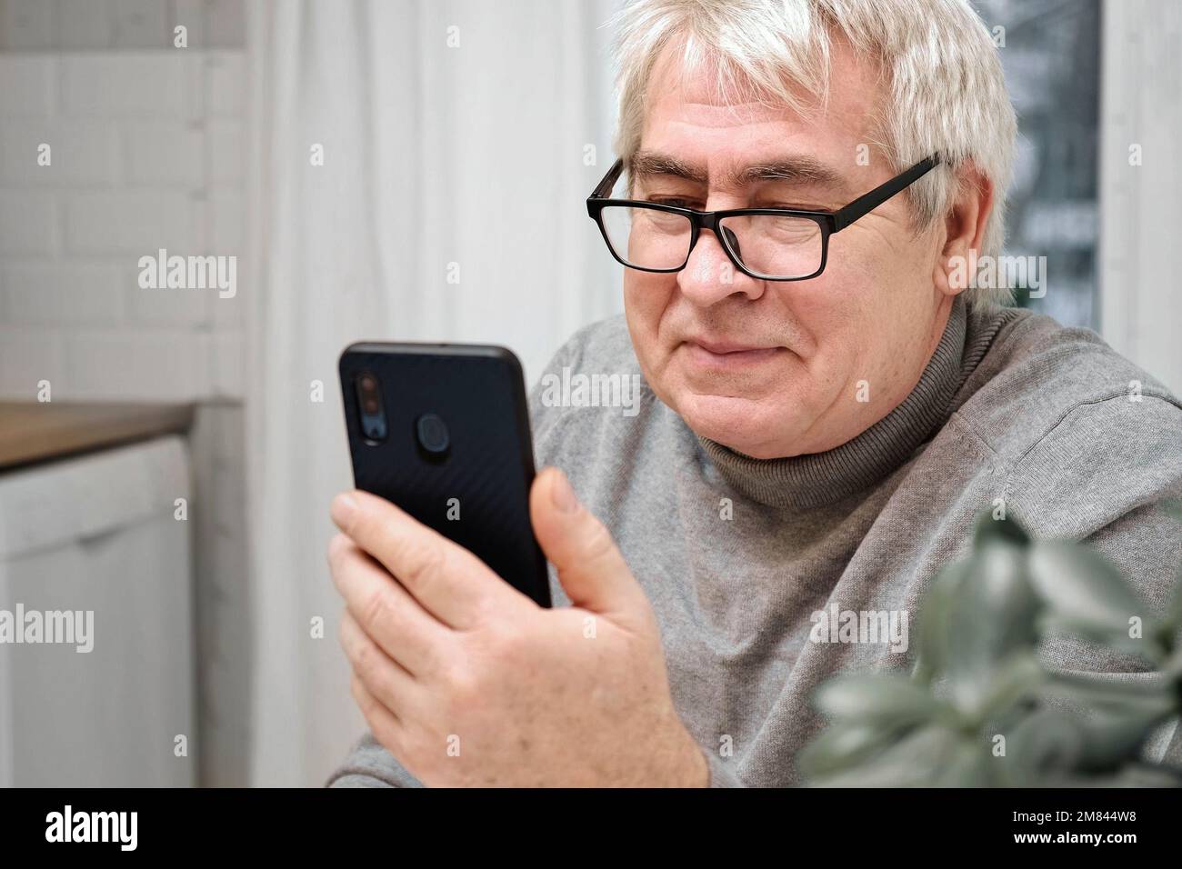 Hoary old man looking at web camera, holding phone, talking with children, wife online. Senior grandpa smiling chatting with friends, family via video Stock Photo