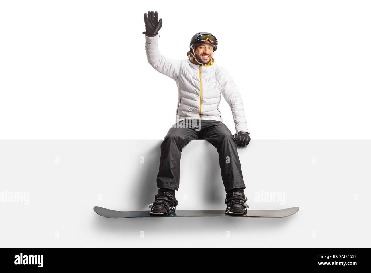Man with a snowboard sitting on a blank panel and waving isolated on white background Stock Photo