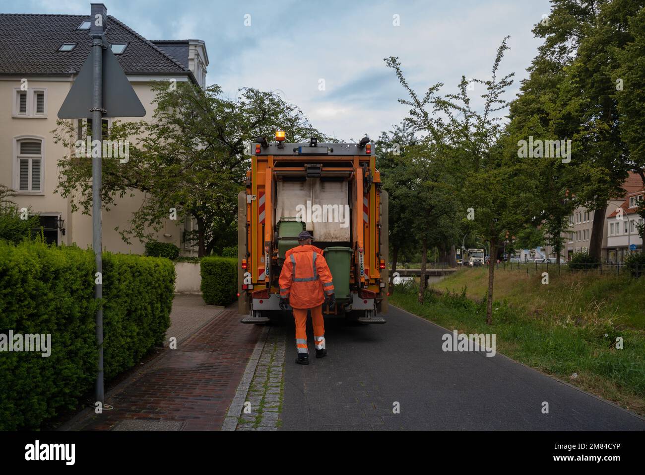 An orange garbage truck stands on a narrow green street, surrounded by