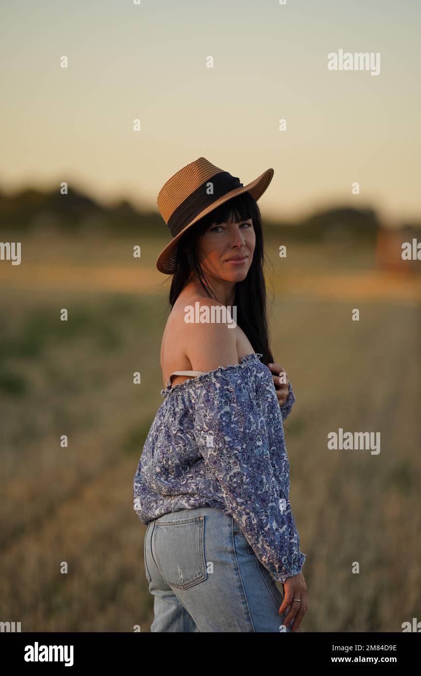 A cowboy woman in a hat and worn jeans stands half a turn on the field and looks into the camera, a country-style woman Stock Photo