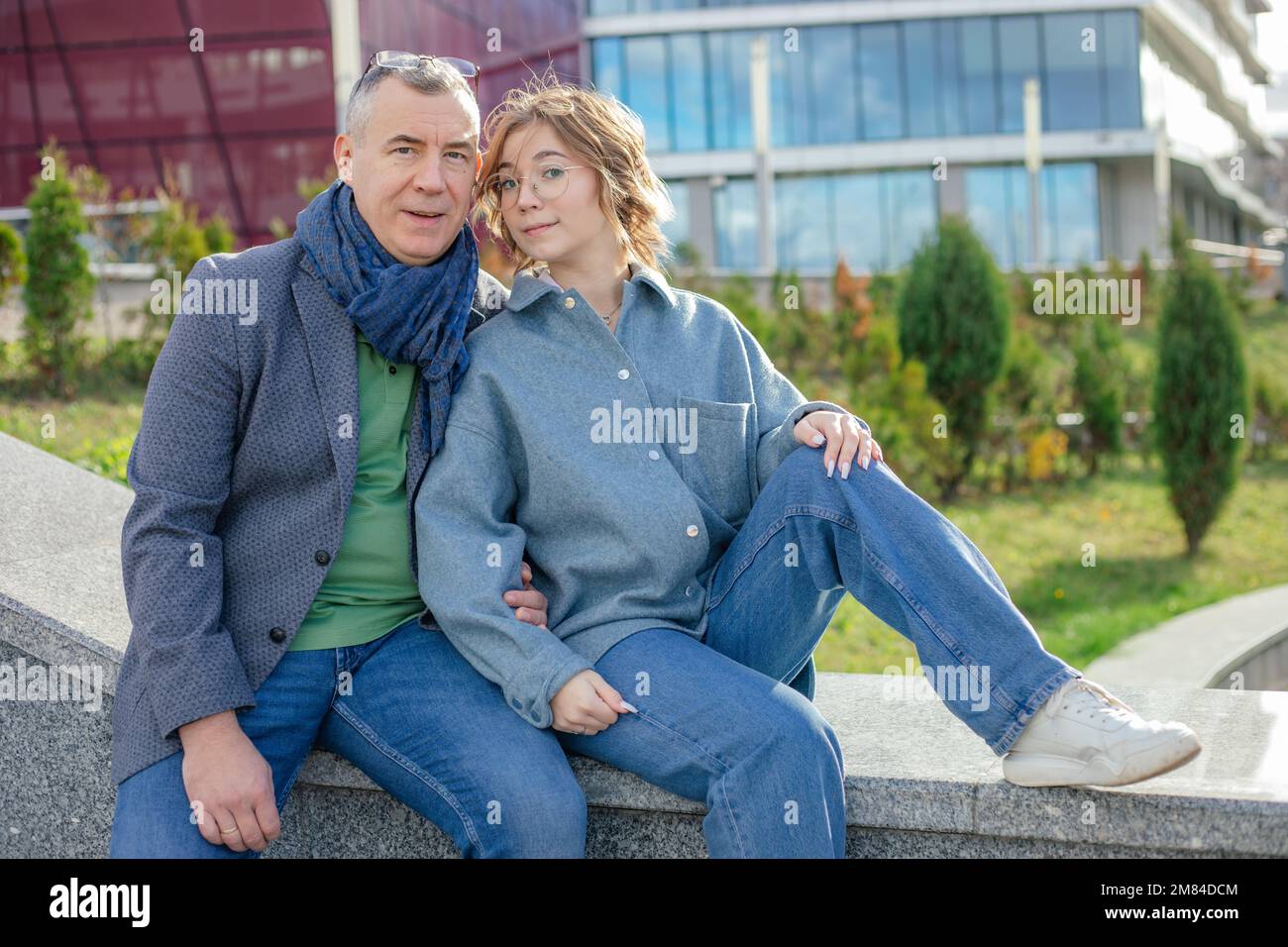 Adult 50s man sit on bench with teenage girl on street near green plants. Father and daughter relax, family walking. Stock Photo