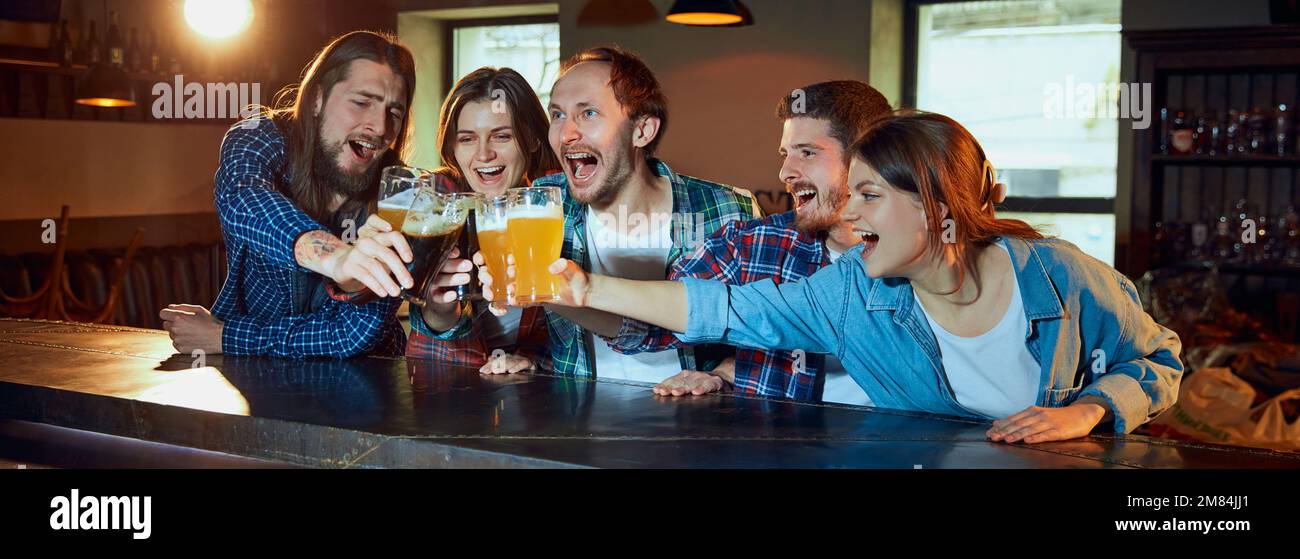Happy young people, friends meeting at pub to watch sport match. Fans emotionally cheering up favourite sport team. Drinking beer. Win Stock Photo
