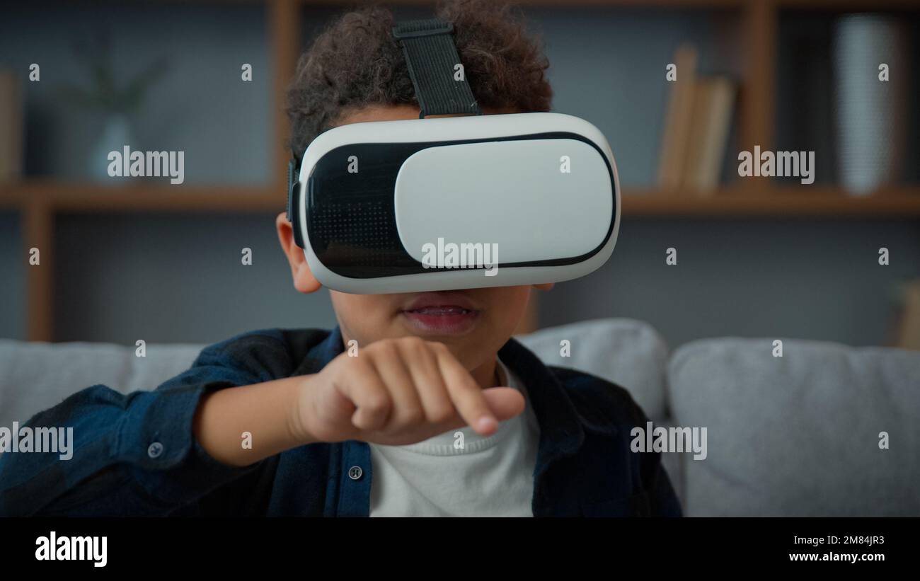 Little African American ethnic boy pupil son kid child schoolboy at home wearing VR glasses play online video 3d game education studying with Stock Photo