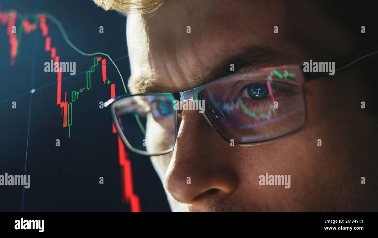 Close-up portrait of focused crypto trader analyst wearing eyeglasses looking at computer screen analyzing stock market charts. Eyeglasses reflection cryptocurrency downtrend charts. Bitcoin crash Stock Photo