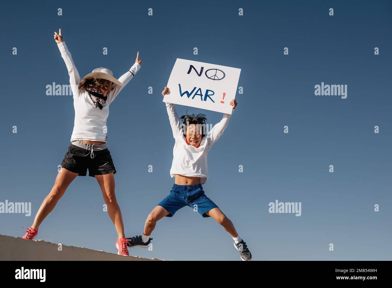 a Caucasian mother in a white hat, jumps next to her Asian son holding a sign that reads 'no war'. concept war and peace. conflict and protest. divers Stock Photo