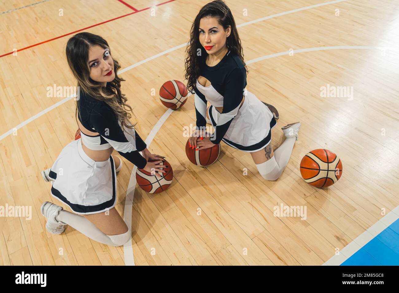 Two vibrant cheerleaders pose in front of the camera, smiling brightly as they hold basketballs on the floor. Dressed in traditional cheer uniforms, complete with mini skirts and knee-high socks. Stock Photo