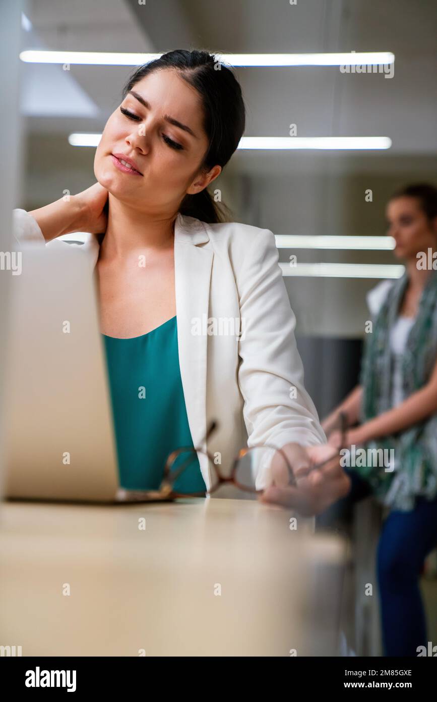 Young woman at office desk working on laptop with stress, neck pain and