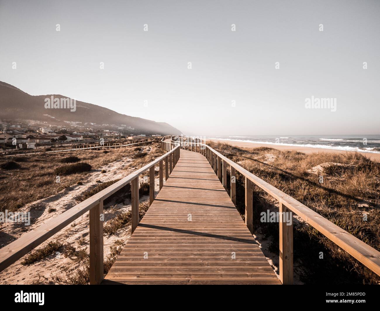 A long boardwalk on a grassy sand dune along the beach leading to the mountain with desaturated colors. The way ahead concept. Quiaios Beach, Portugal Stock Photo