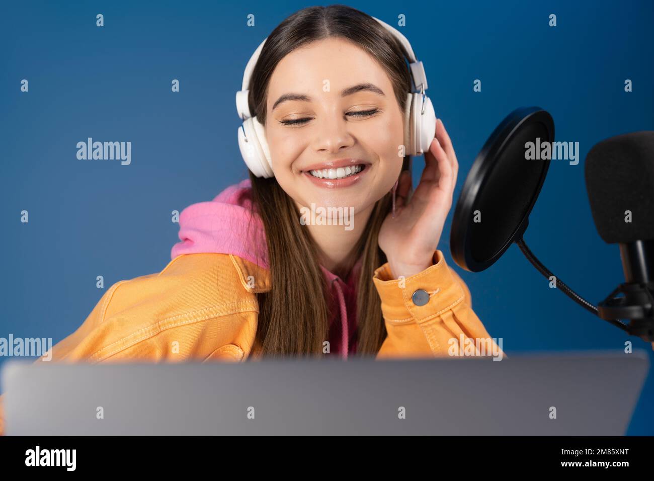 Pleased teenager in headphones looking at laptop near microphone isolated on blue Stock Photo