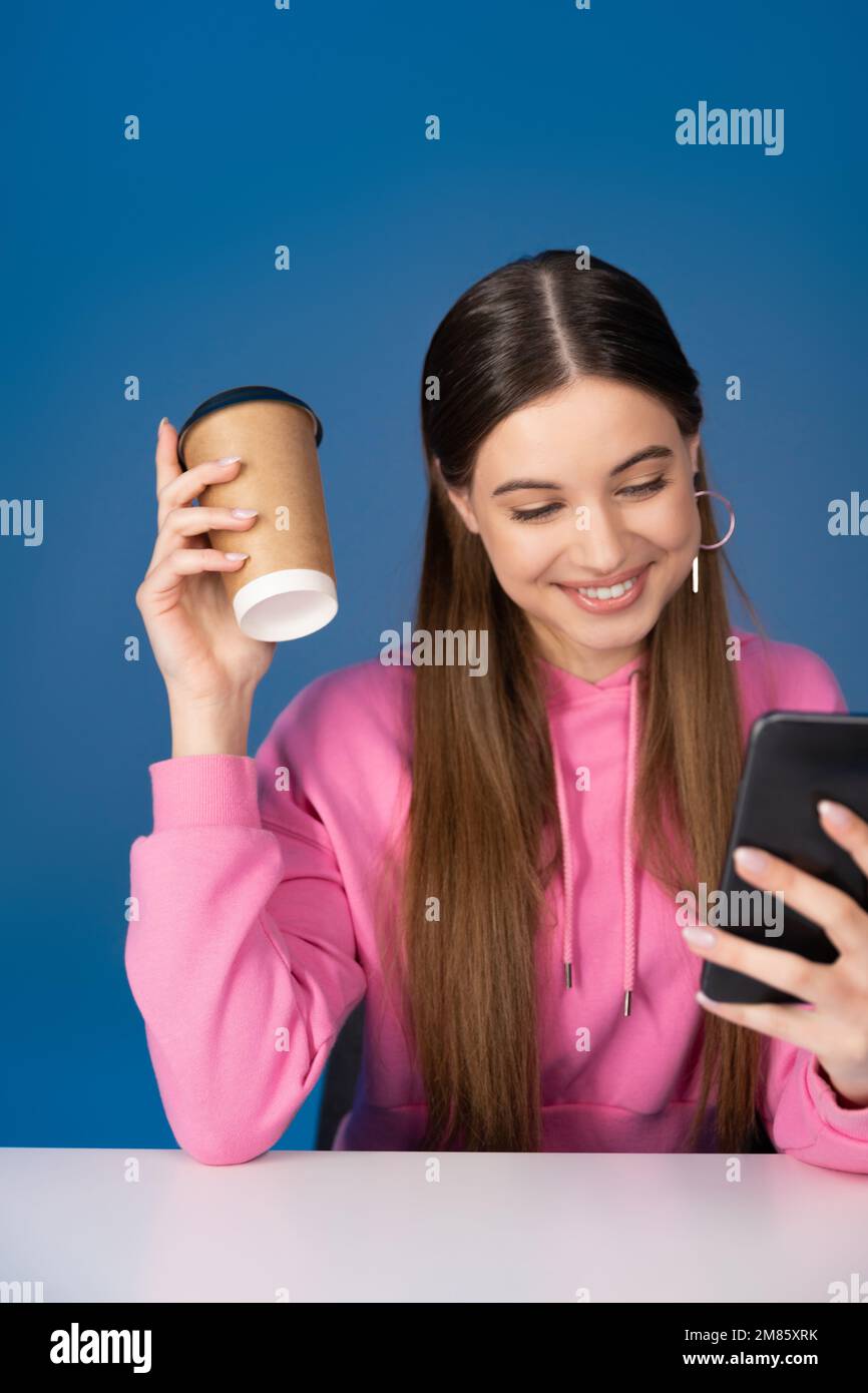 Positive teen girl holding coffee to go and using smartphone isolated on blue Stock Photo