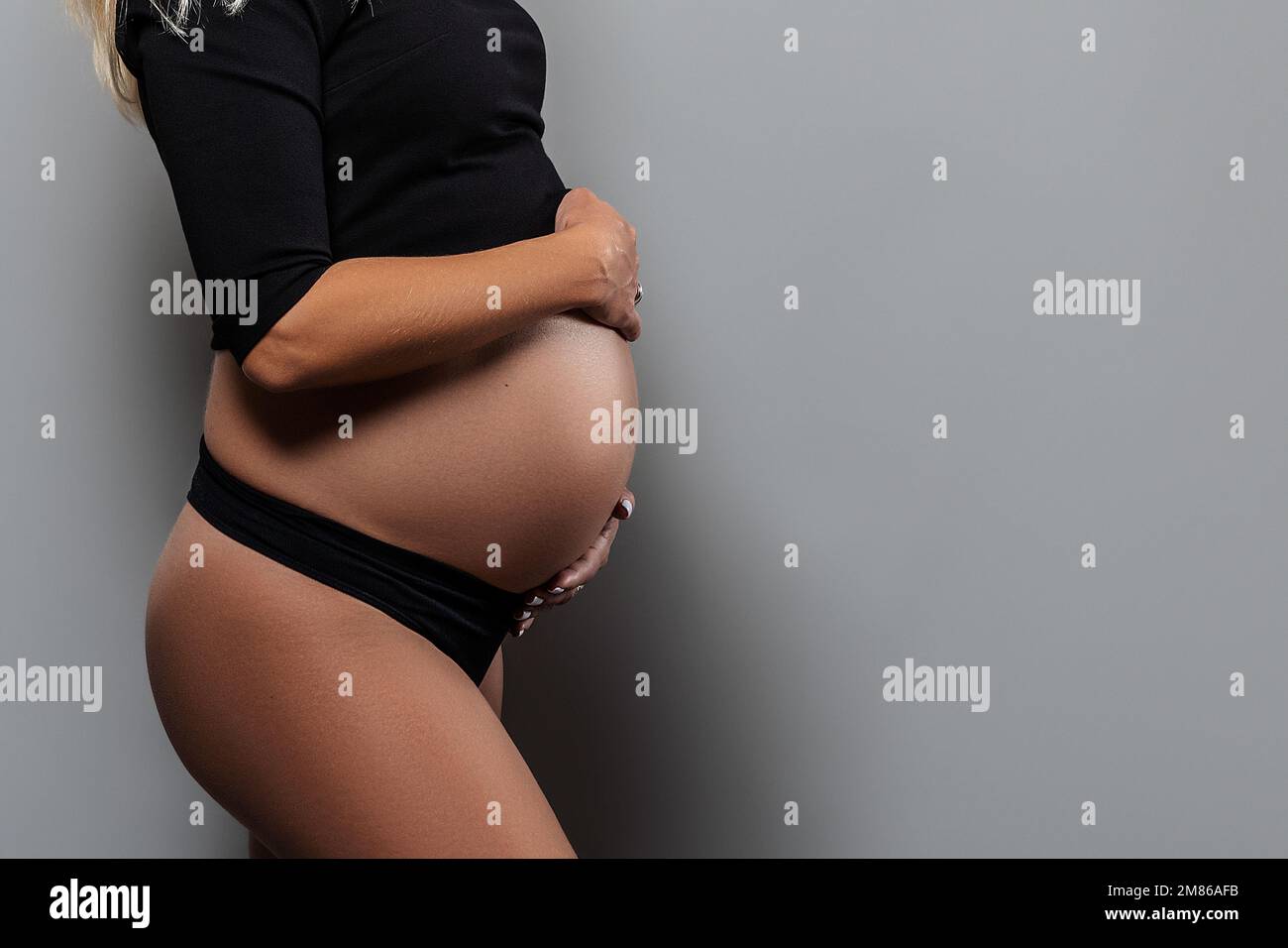 A pregnant woman holds her hands on her stomach on a gray background. Pregnancy, maternity, preparation and expectation concept. Stock Photo