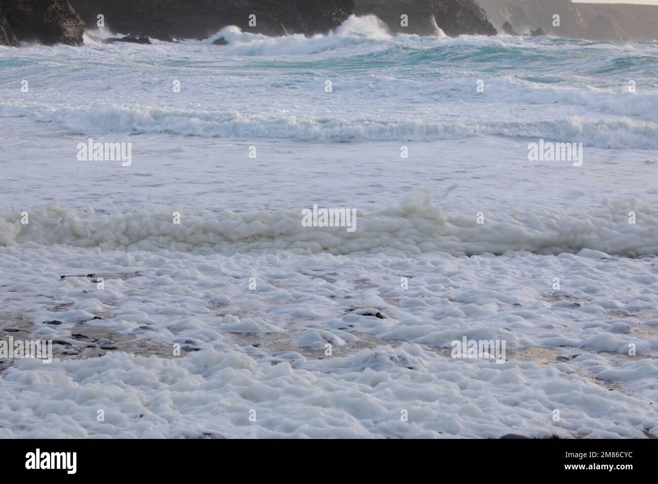 Seafoam on a Cornish beach with crashing waves in the background.  Dollar Cove, The Lizard, Cornwall Stock Photo