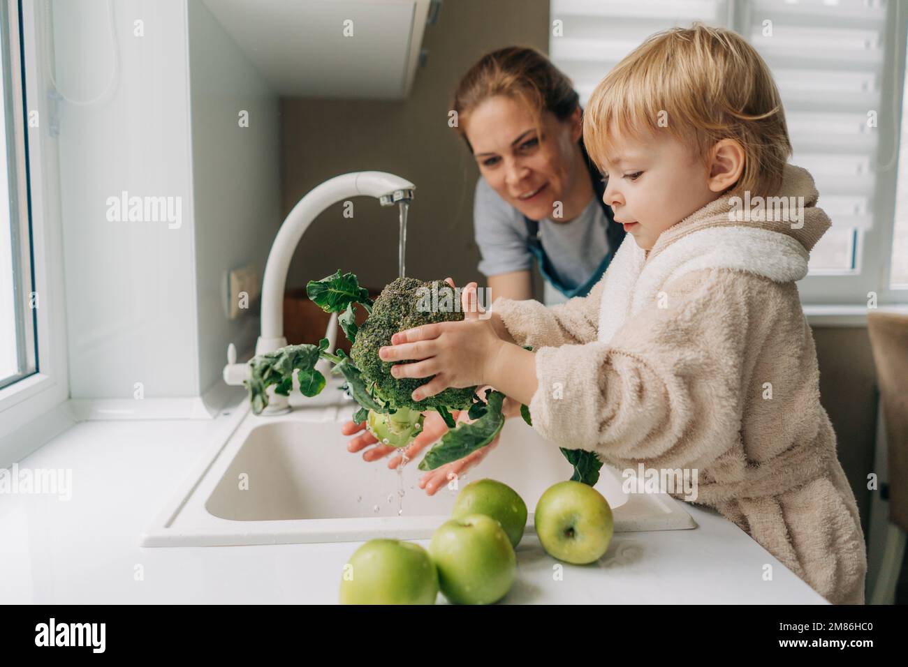 A mother and a small child are washing broccoli together in the kitchen for preparing dinner. Stock Photo