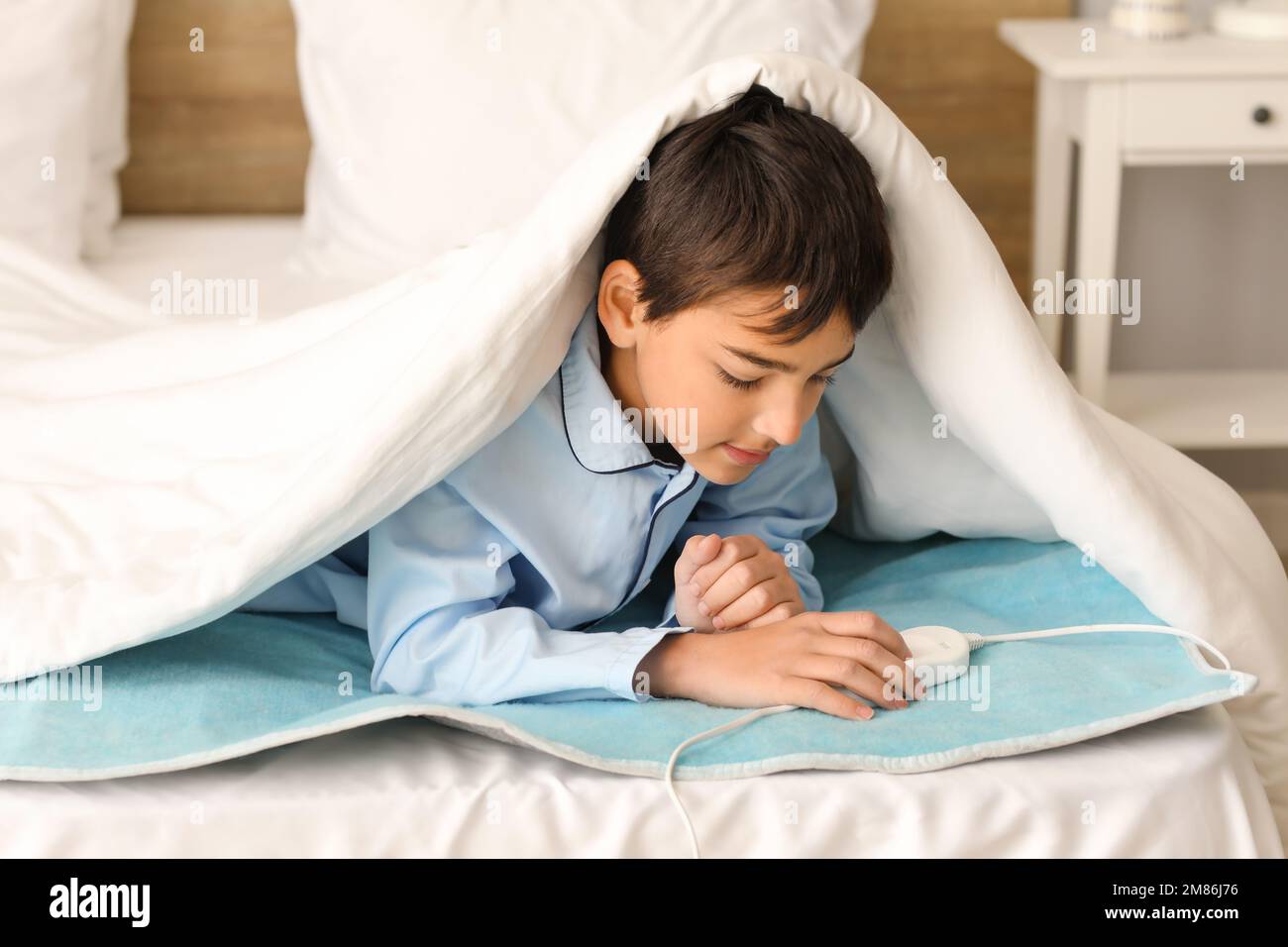 Little boy lying under blanket on electric heating pad in bedroom Stock