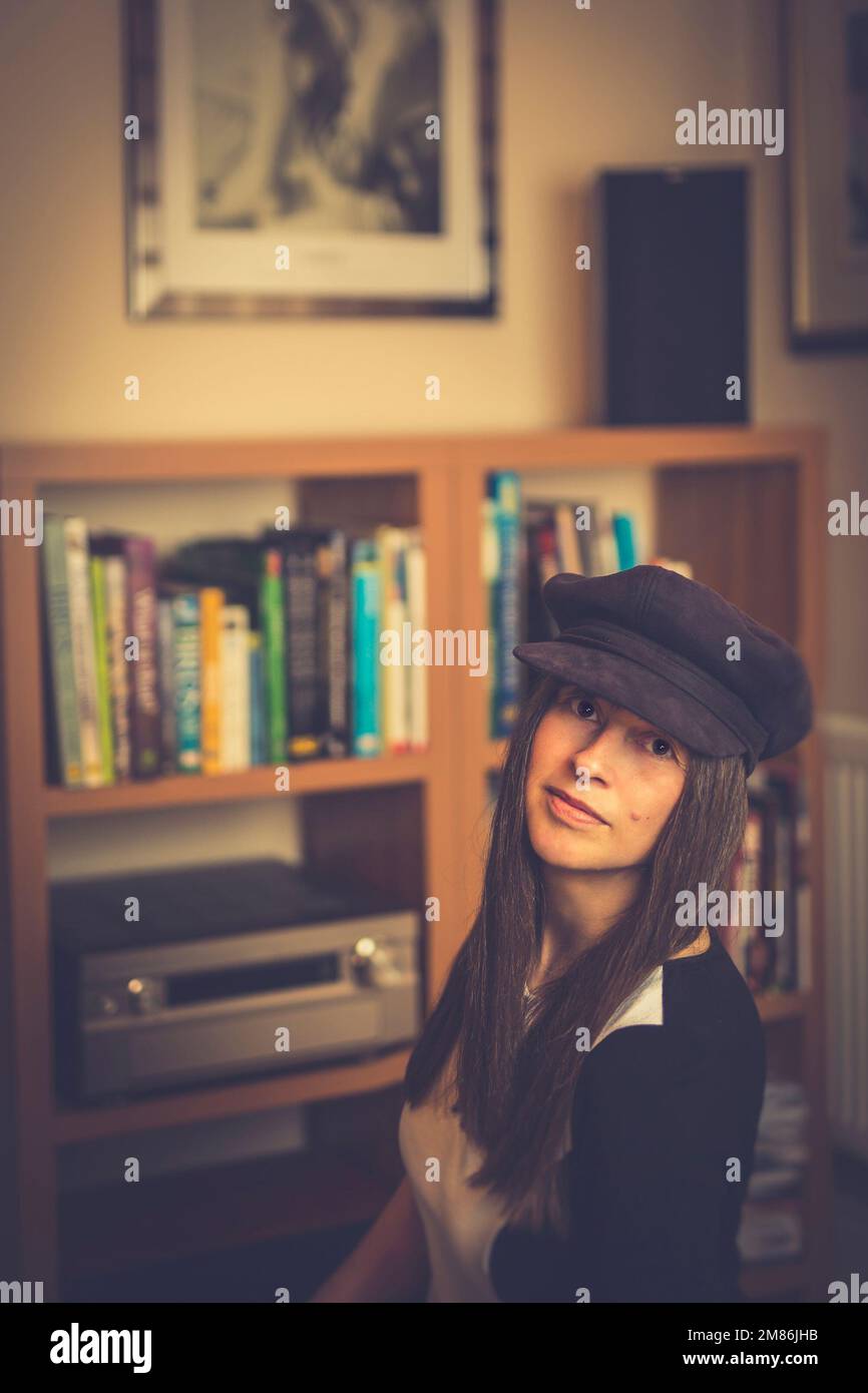 British female, with long hair & wearing 1970s style hat, posing for photograph isolated indoors at home in a typical UK lounge/living room. Stock Photo