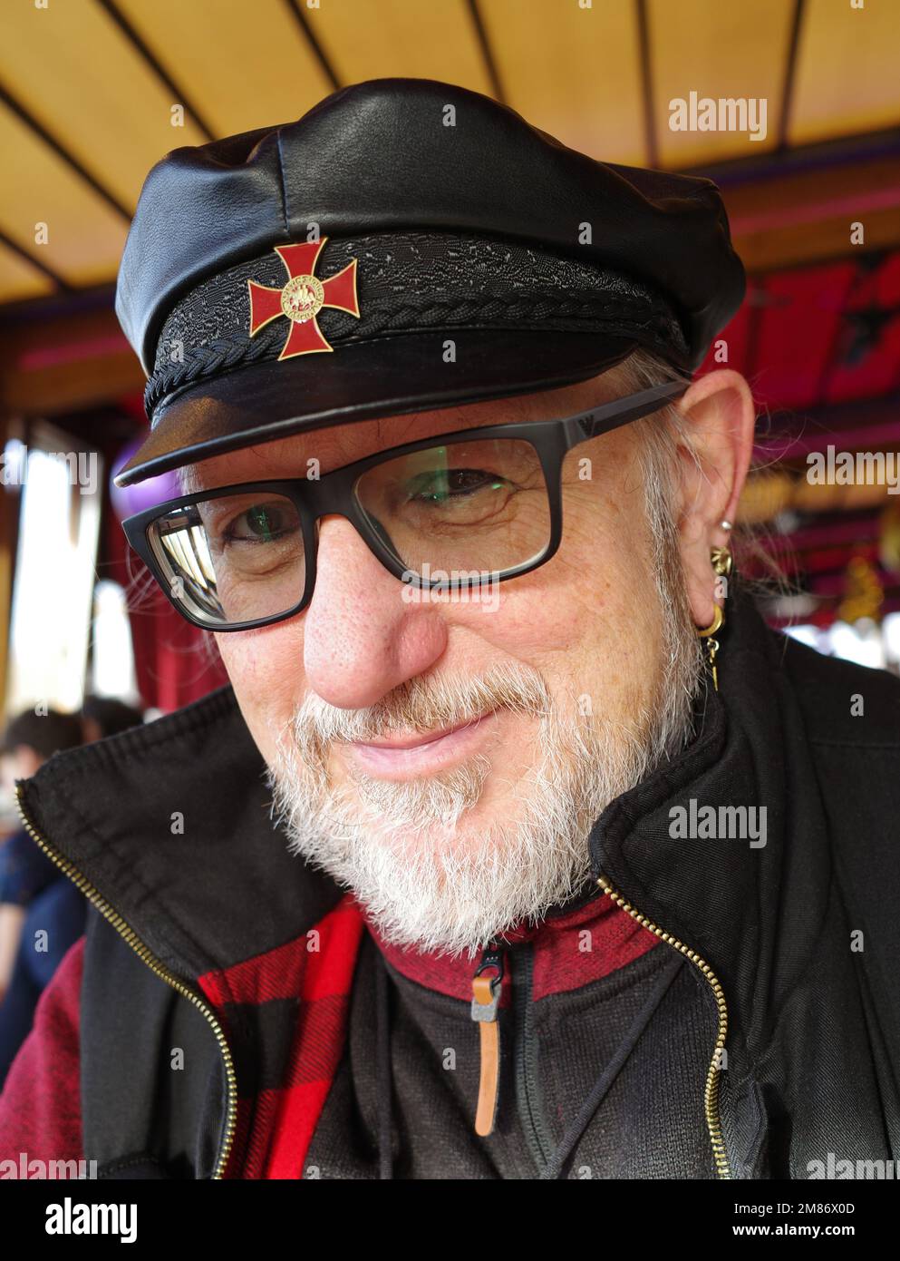 Portrait of friendly looking man of almost seventy with glasses, beard, earrings and a captains cap with Templar cross Stock Photo