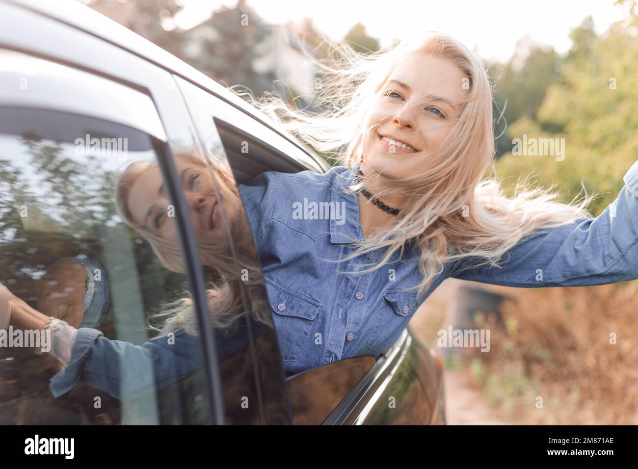 Dreaming woman joyfully leave city on sunny day sticking head out of open car window and looking up to sky. Young blonde lady with happy mood go to Stock Photo