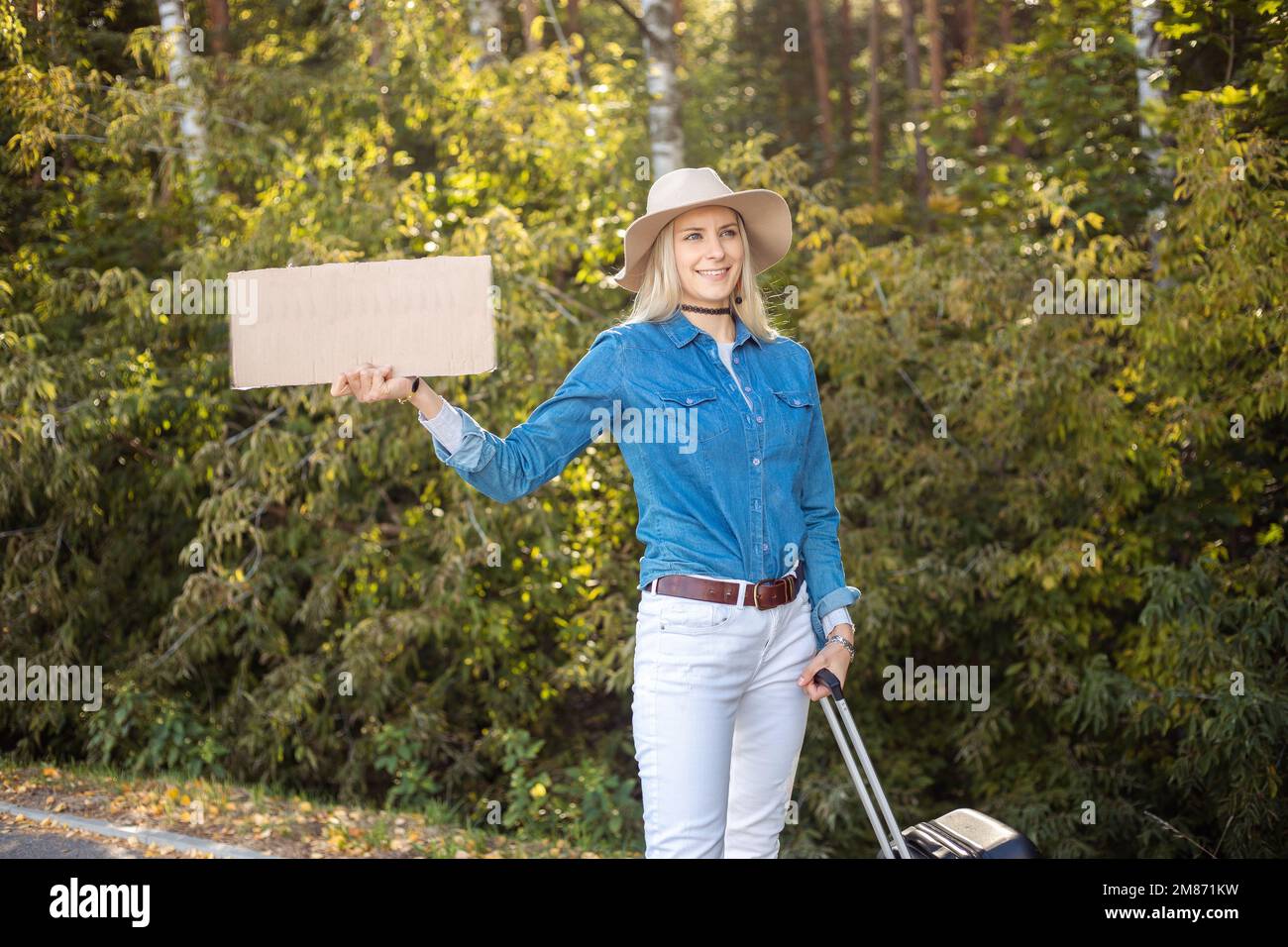 Happy woman wait passing car in forest standing with suitcase and cardboard poster on roadside. Blonde lady escape from city to go anywhere Stock Photo
