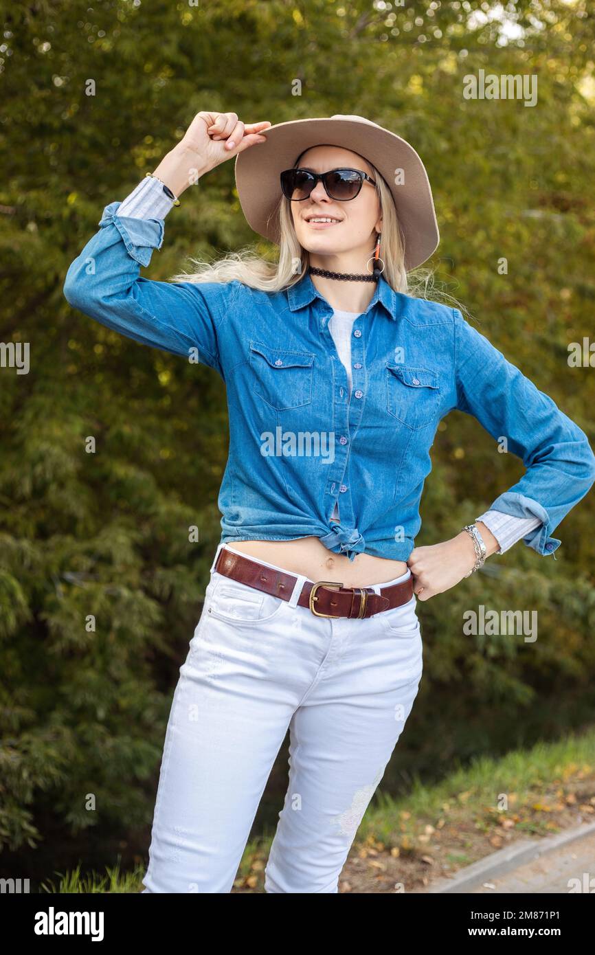 Portrait of dreaming blonde woman in sunglasses and elegant hat standing in forest on roadside. Happy traveller in seasonal denim outfit. Autumn Stock Photo