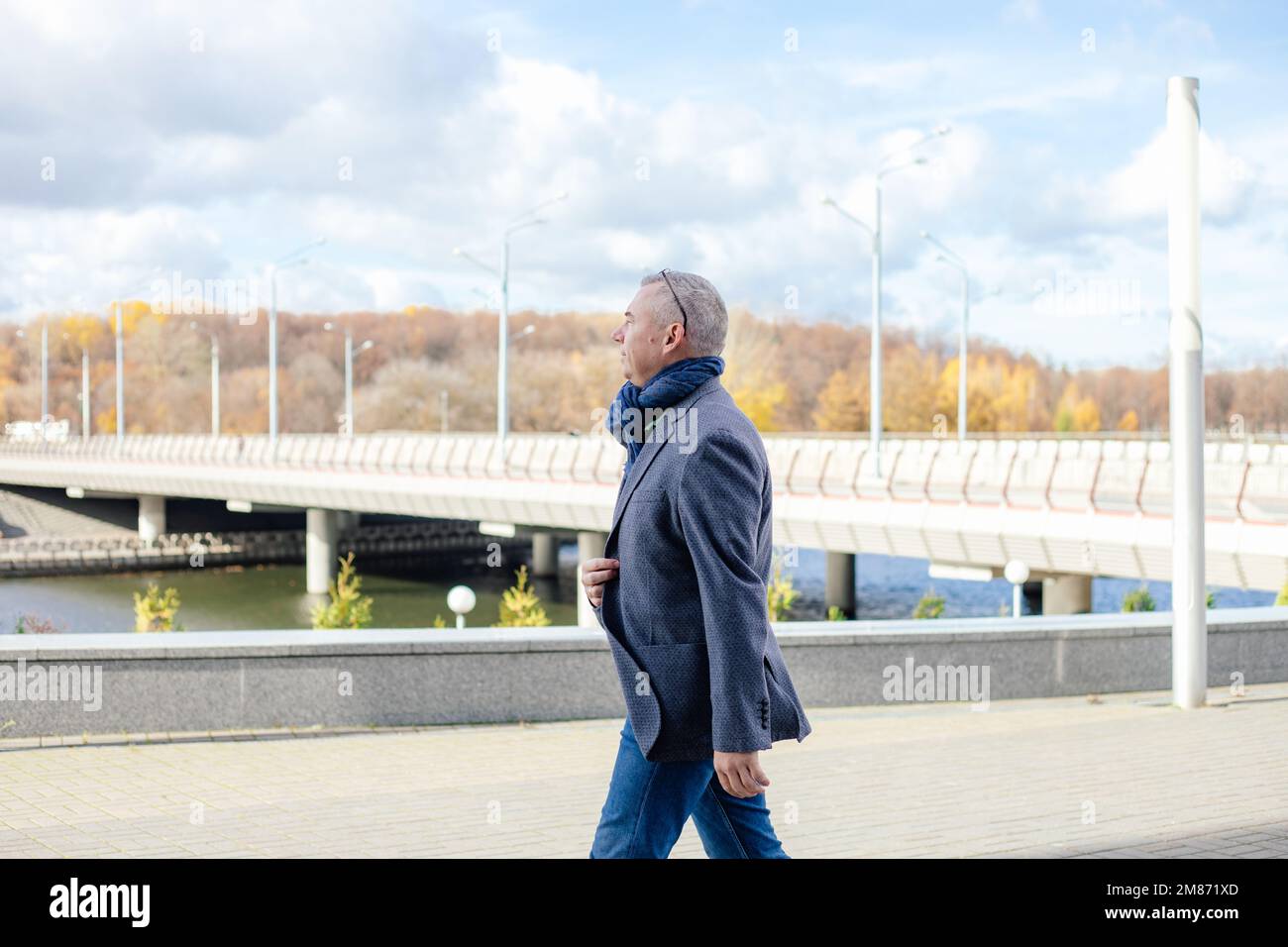 Calm gray haired man looking away camera in warm jacket and scarf walking street, near promenade bridge. Low temperature Stock Photo