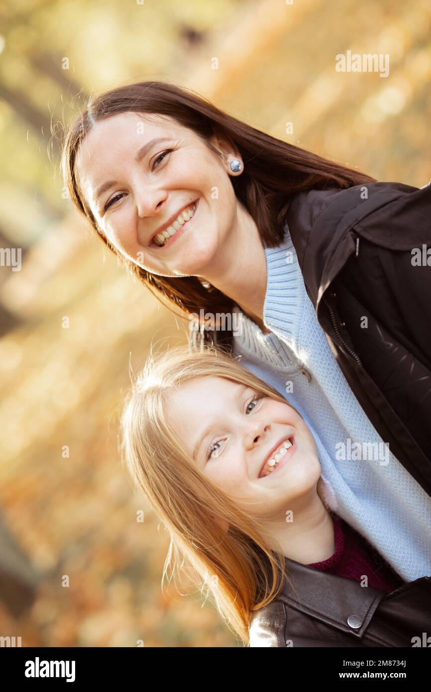 Vertical cheerful, glad, laughing family, mother and little daughter look at camera, stroking and stand in autumn park Stock Photo