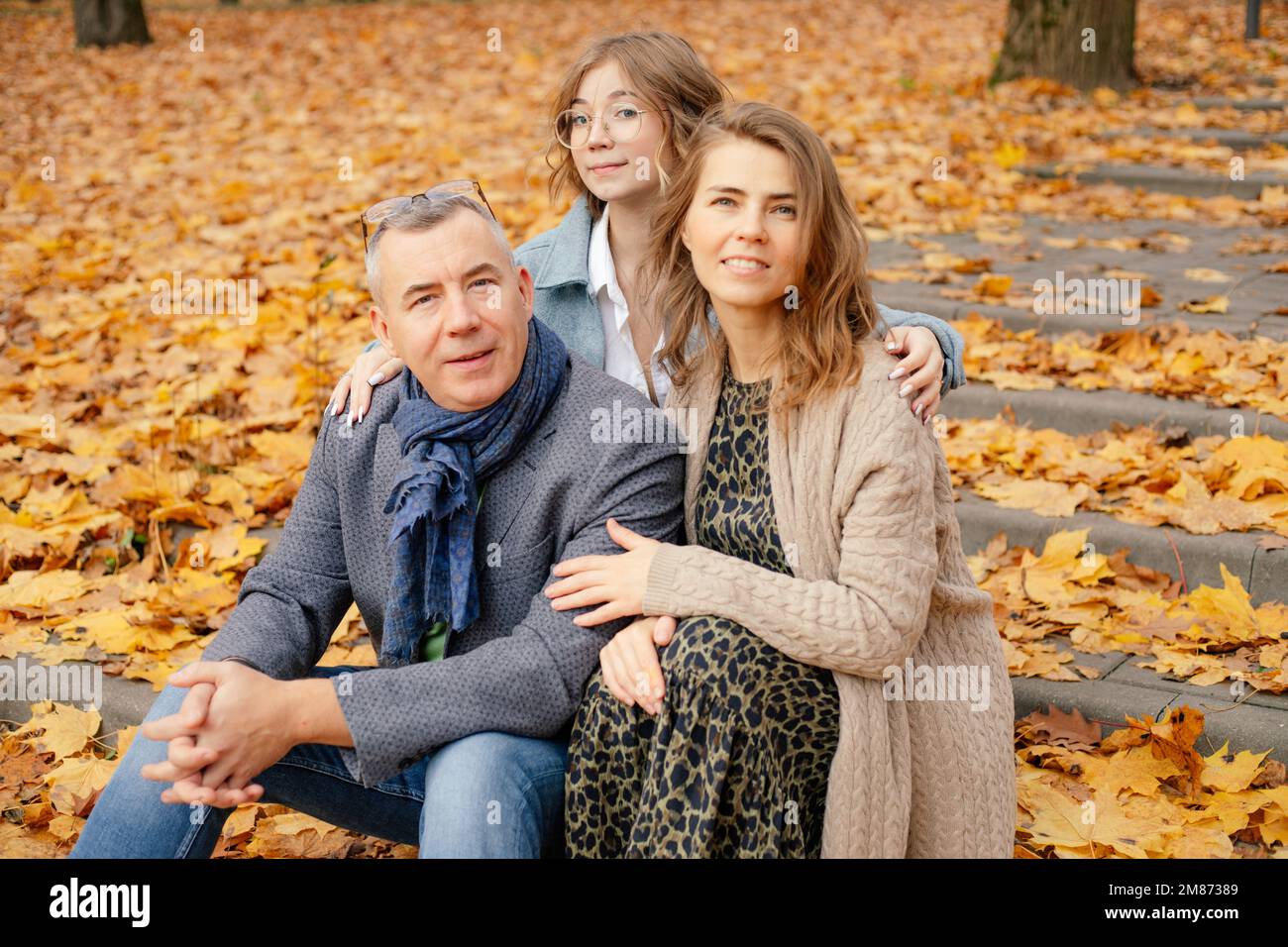 Merry, smiling, curious family of mother, father and daughter hugging together, sit on steps, relax. Family traditions Stock Photo