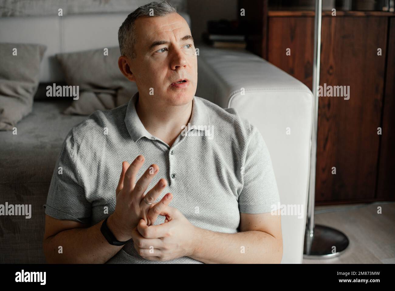 Nervous tired dispelled mature grizzled man looking out window, hold and rub hand, sit near sofa indoor. Tunnel syndrome Stock Photo